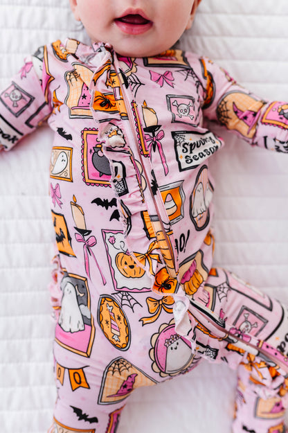 A baby wearing a pink footed sleeper with a colorful Halloween print, lying on a white quilted blanket. The print features pumpkins, ghosts, bats, and the text 'Spooky Season'.