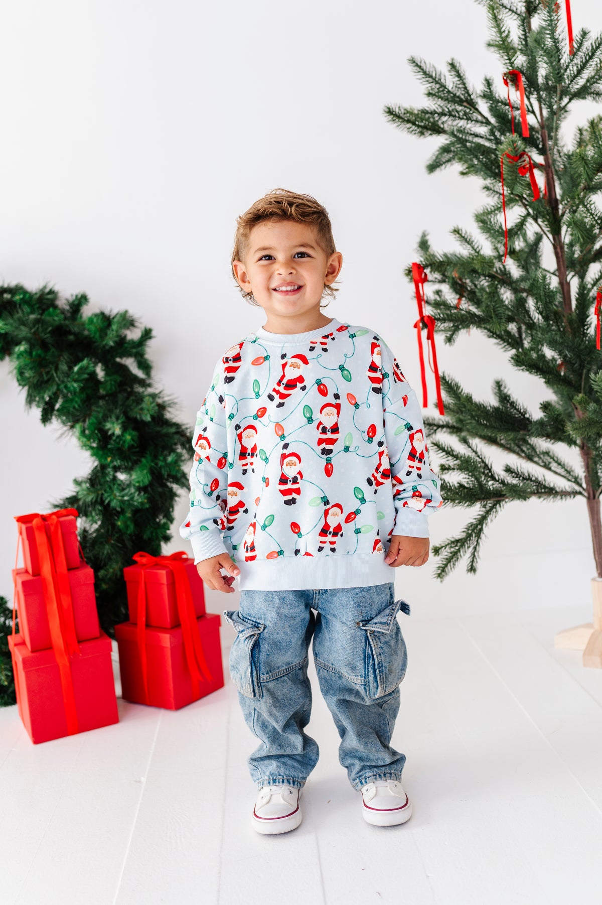 A young boy smiling and wearing a light blue long-sleeve crewneck sweatshirt with a festive Santa Claus and candy cane print, paired with blue jeans and white sneakers. He is standing in a bright room decorated with a Christmas tree, red ornaments, and wrapped red presents.