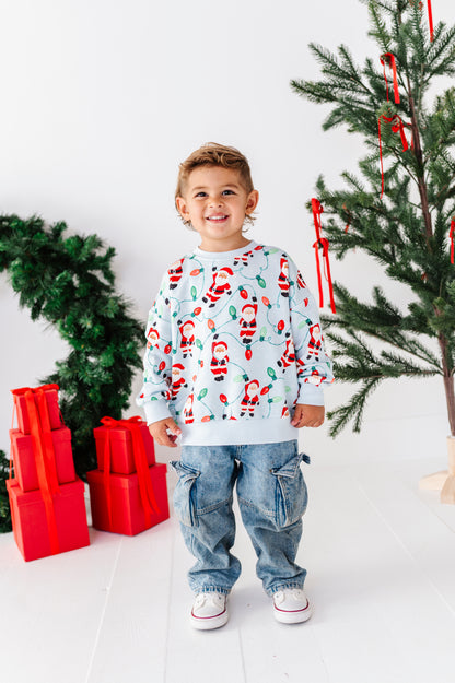A young boy smiling and wearing a light blue long-sleeve crewneck sweatshirt with a festive Santa Claus and candy cane print, paired with blue jeans and white sneakers. He is standing in a bright room decorated with a Christmas tree, red ornaments, and wrapped red presents.