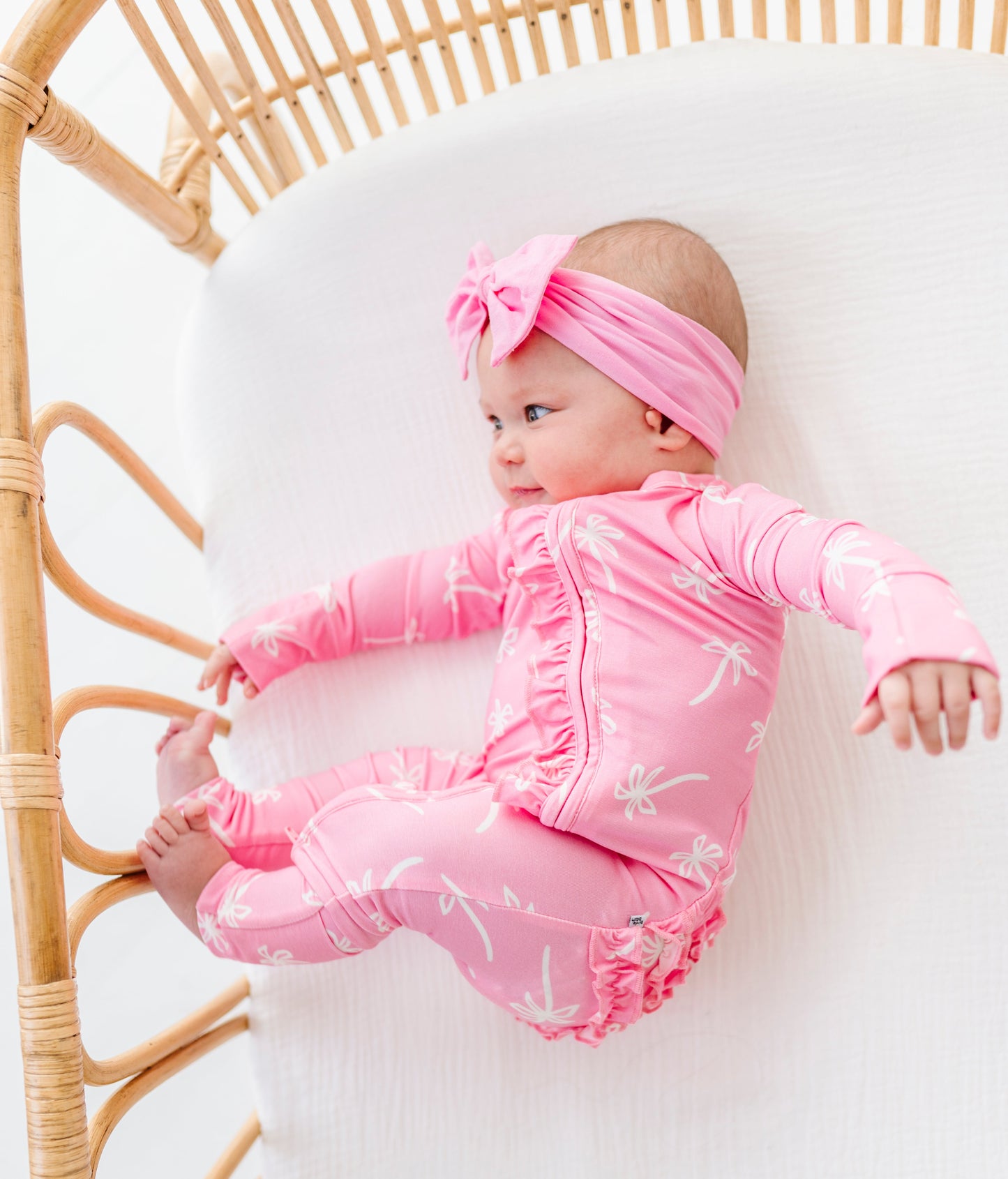 Baby in pink outfit lying on a white blanket with a wicker bassinet.