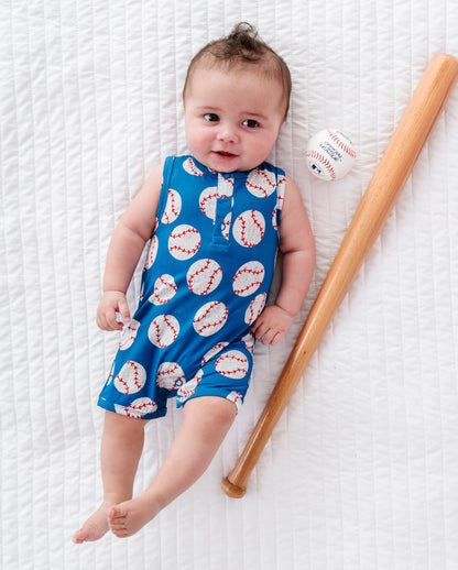 Baby in a blue romper with baseball pattern lying on a white textured surface next to a wooden bat and small ball.