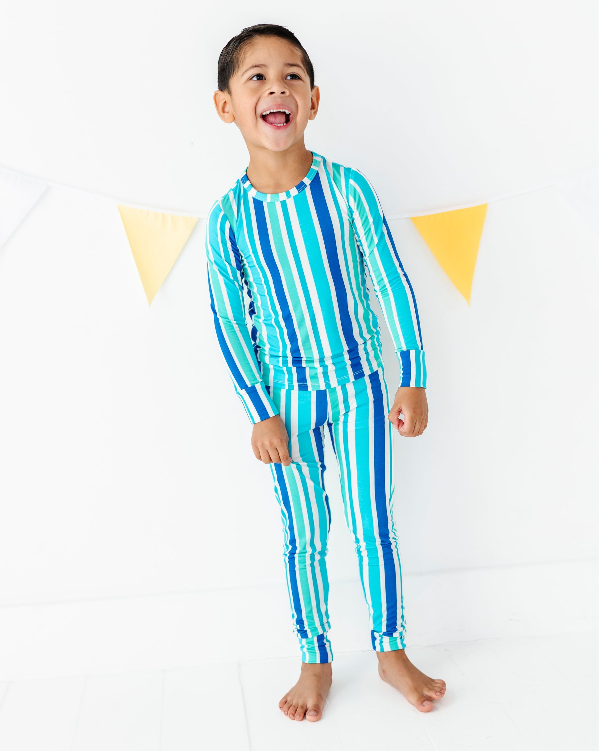 A young boy smiling and laughing while wearing a matching long-sleeve striped swim shirt and swim pants set in shades of blue, teal, and white, standing against a white studio background with yellow bunting.