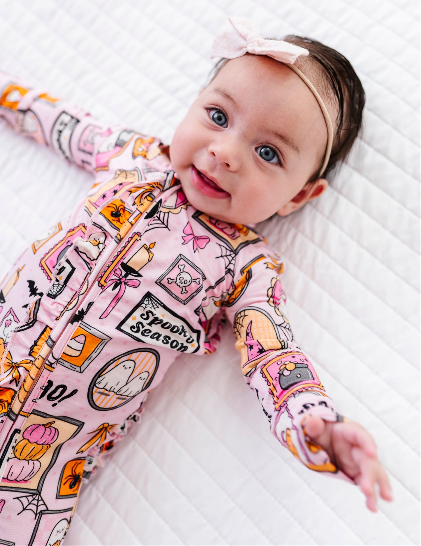 A baby lying on a white quilted mattress, wearing a pink long-sleeve pajama top with a colorful 'Spooky Season' graphic print and a matching pink bow headband.