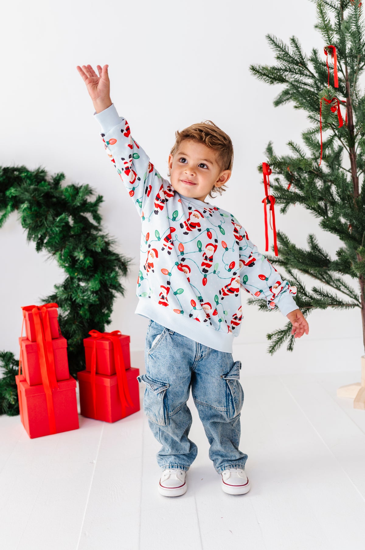 A young boy wearing a light blue Christmas sweatshirt with a Santa Claus pattern, blue jeans, and white sneakers, standing in a festive room with a Christmas tree, red ornaments, and wrapped presents.