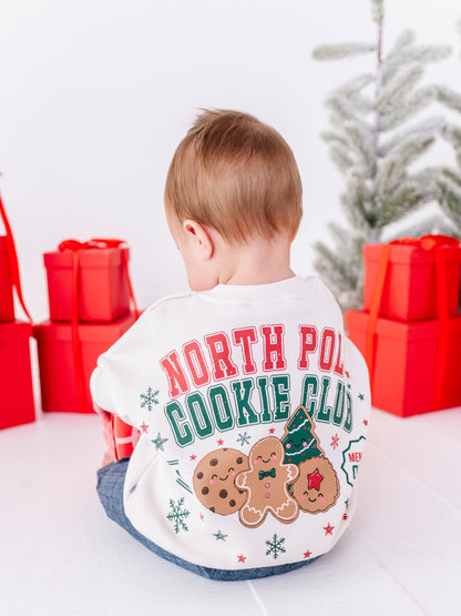 A baby boy playing in front of a Christmas tree, wearing a white crewneck that says "NORTH POLE COOKIE CLUB" across the back with gingerbread cookies 