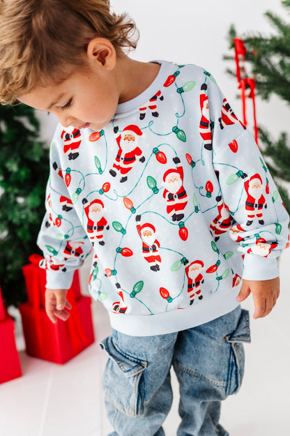 A young child wearing a white long-sleeve crewneck sweatshirt with a festive Christmas print of Santa Claus and holiday lights, paired with blue jeans, standing in front of a decorated Christmas tree and wrapped gifts.