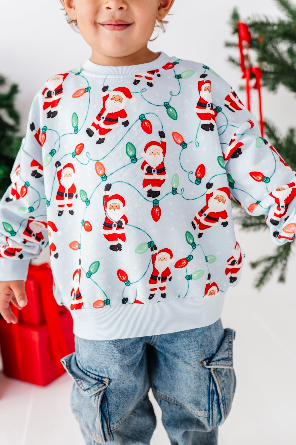 A child wearing a white crewneck sweatshirt with a festive Christmas print of Santa Claus and colorful holiday lights, paired with blue jeans.