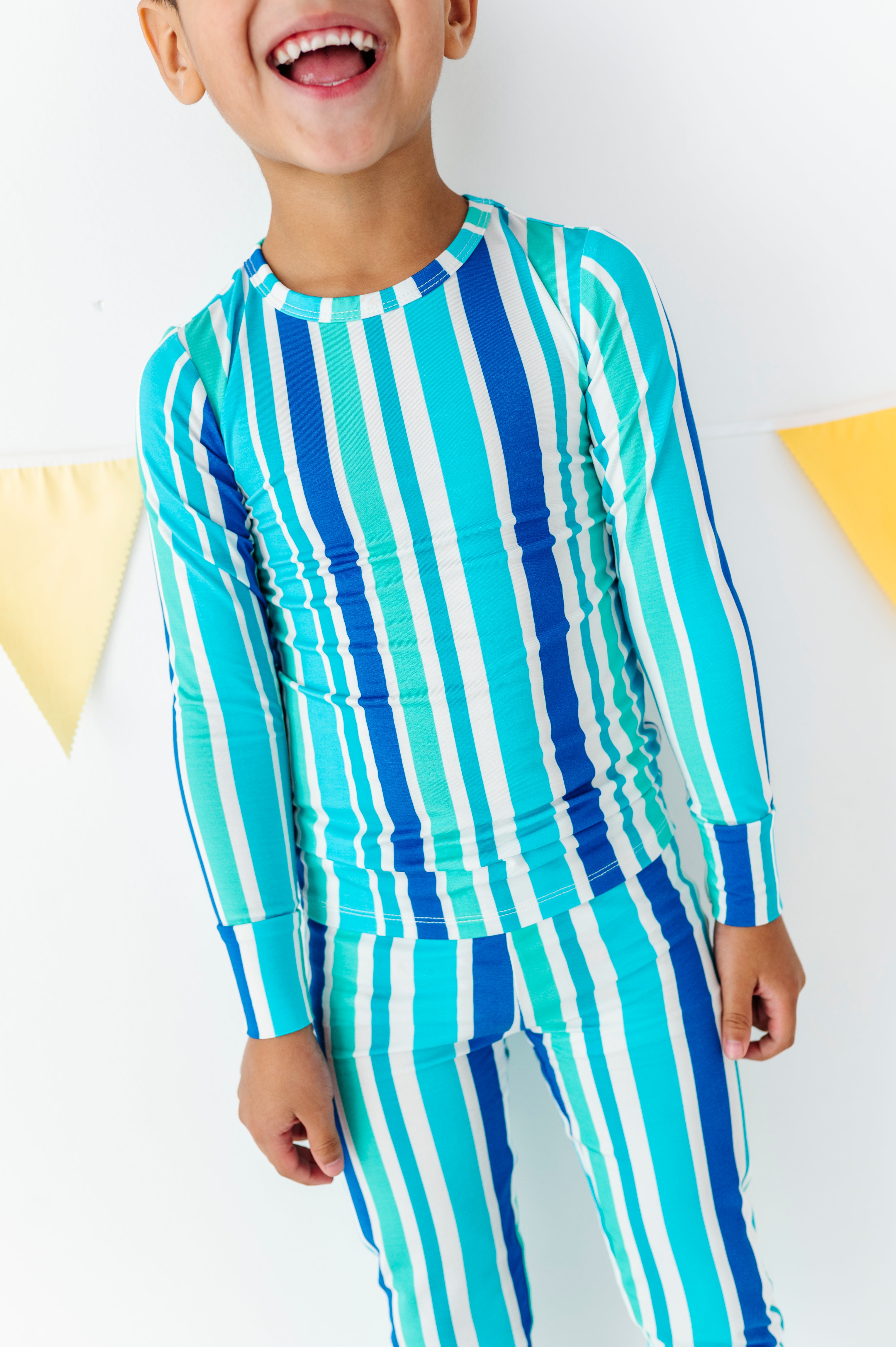 A child wearing a matching long-sleeve top and pants set with vertical stripes in turquoise, blue, and white, standing against a white wall with yellow bunting.