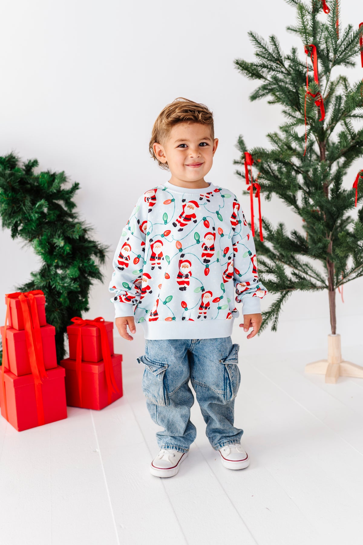 A young boy stands in a bright studio setting wearing a light blue long-sleeve crewneck sweatshirt with a red and green Santa Claus pattern, paired with blue jeans and white sneakers. Christmas trees and red wrapped gifts are visible in the background.