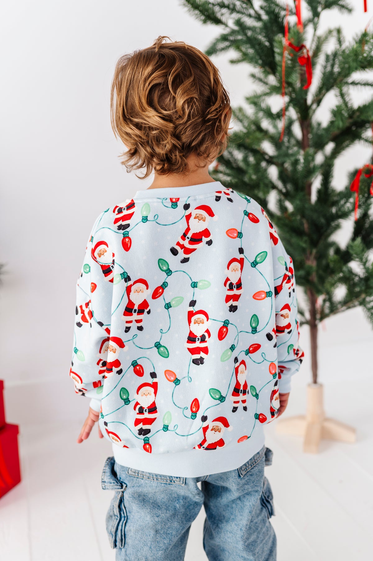 A child viewed from behind wearing a light blue long-sleeve sweatshirt with a festive Christmas print of Santa Claus figures and red, green, and white holiday lights, paired with blue jeans in a bright indoor setting with a Christmas tree in the background.