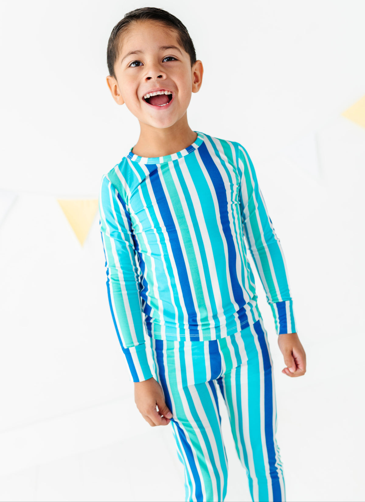 A young boy smiling and wearing a matching two-piece swimsuit with vertical stripes in turquoise, blue, and white, set against a clean white background with subtle yellow geometric accents.