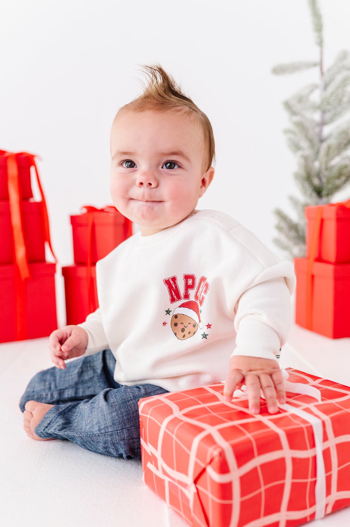 A baby boy playing with a red wrapped present wearing denim jeans and a white crewneck sweatshirt with the letters "NPCC" and a chocolate chip cookie on the chest 