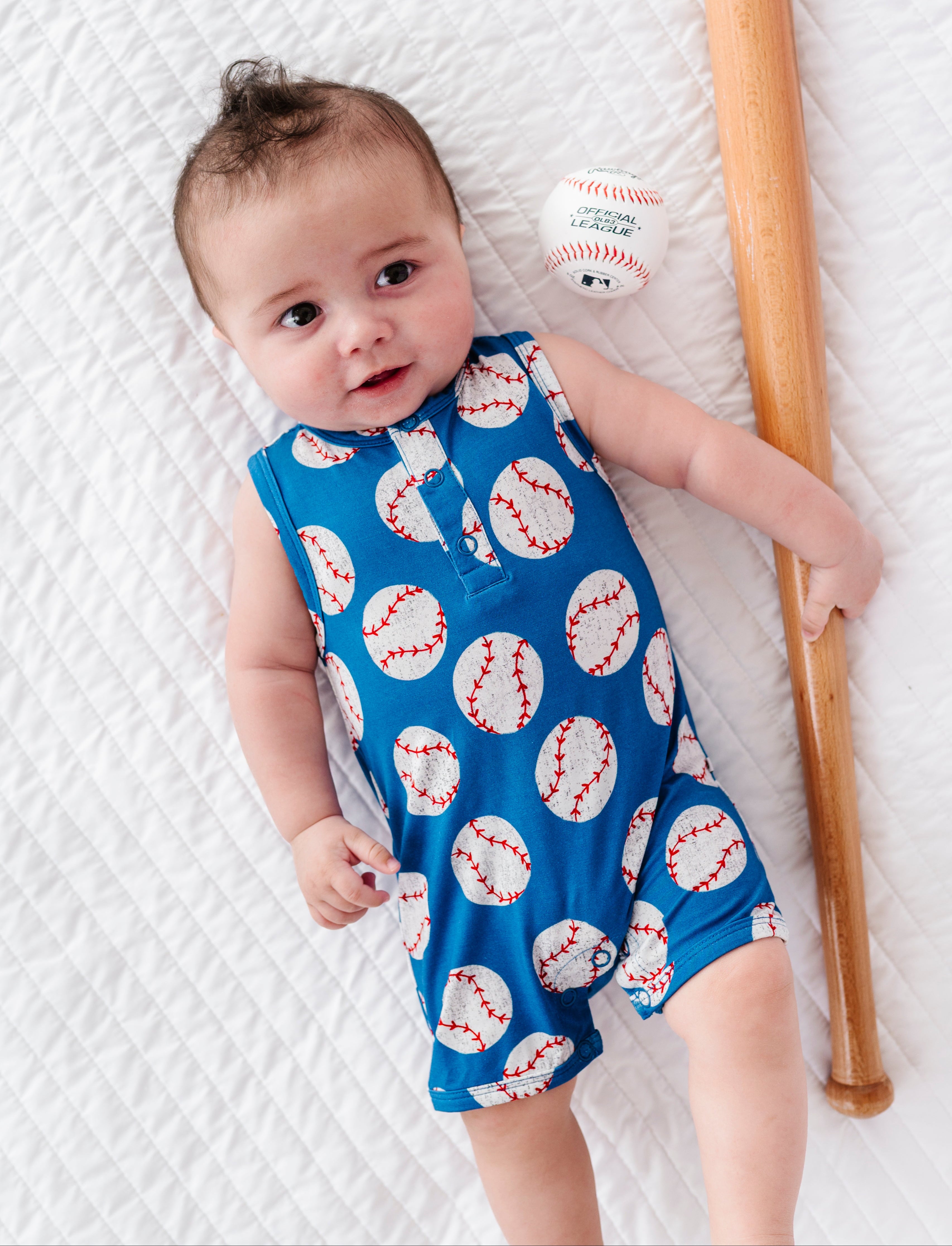 Baby wearing a blue bamboo romper with baseball pattern, holding a baseball and bat on a white textured surface