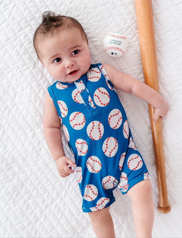 Baby wearing a blue bamboo romper with baseball pattern, holding a baseball and bat on a white textured surface