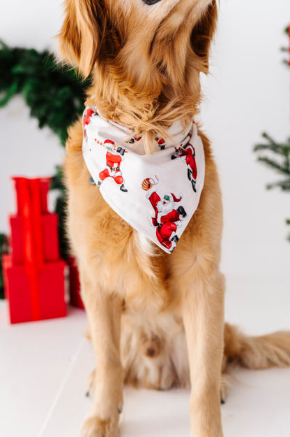 A golden retriever dog wearing a white bandana with a Santa Claus print, sitting in a festive indoor setting with Christmas decorations in the background.