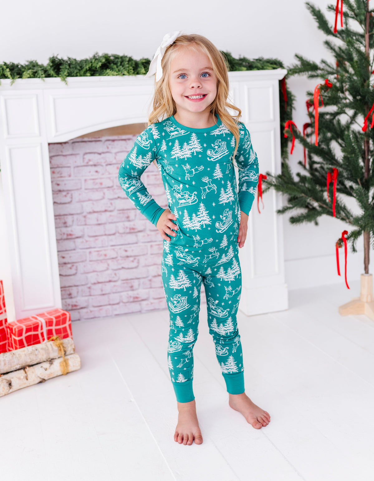 A blonde little girl wearing a bamboo two-piece pajama set. The pajamas feature vintage sleighs, Santas, reindeer, and pine trees. She's standing in front of a fireplace and a ribbon-filled Christmas tree