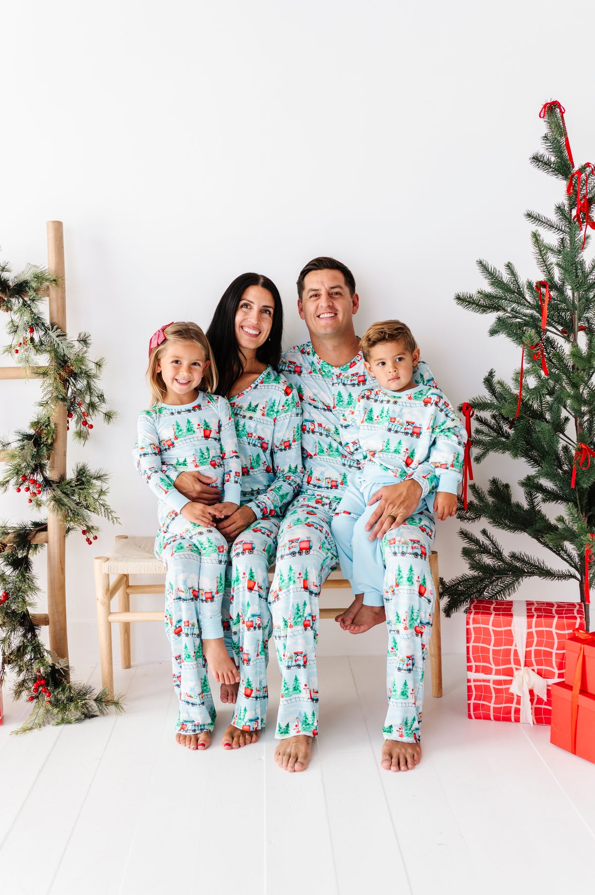 A family of four wearing matching Christmas-themed bamboo pajamas sits together on a white bench in a bright, holiday-decorated room. The pajamas feature a light blue background with a repeating pattern of green trees, red ornaments, and small red vehicles.