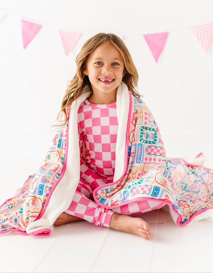 Young girl wrapped in a colorful blanket with checkered pajamas, sitting on a white floor with pink triangle flags in the background.