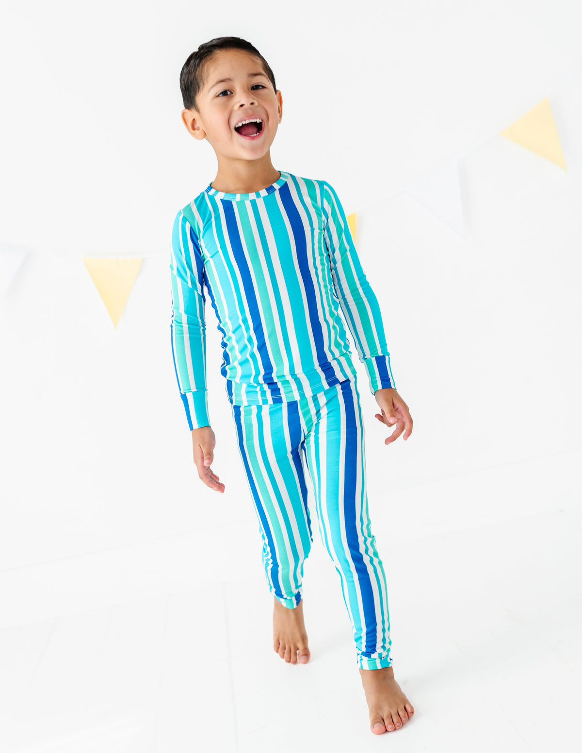 A young boy wearing a matching two-piece long-sleeve pajama set with vertical stripes in shades of blue, teal, and white, standing against a white studio background with subtle yellow triangle decorations.