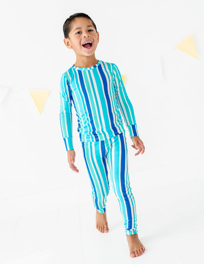 A young boy wearing a matching two-piece long-sleeve pajama set with vertical stripes in shades of blue, teal, and white, standing against a white studio background with subtle yellow triangle decorations.