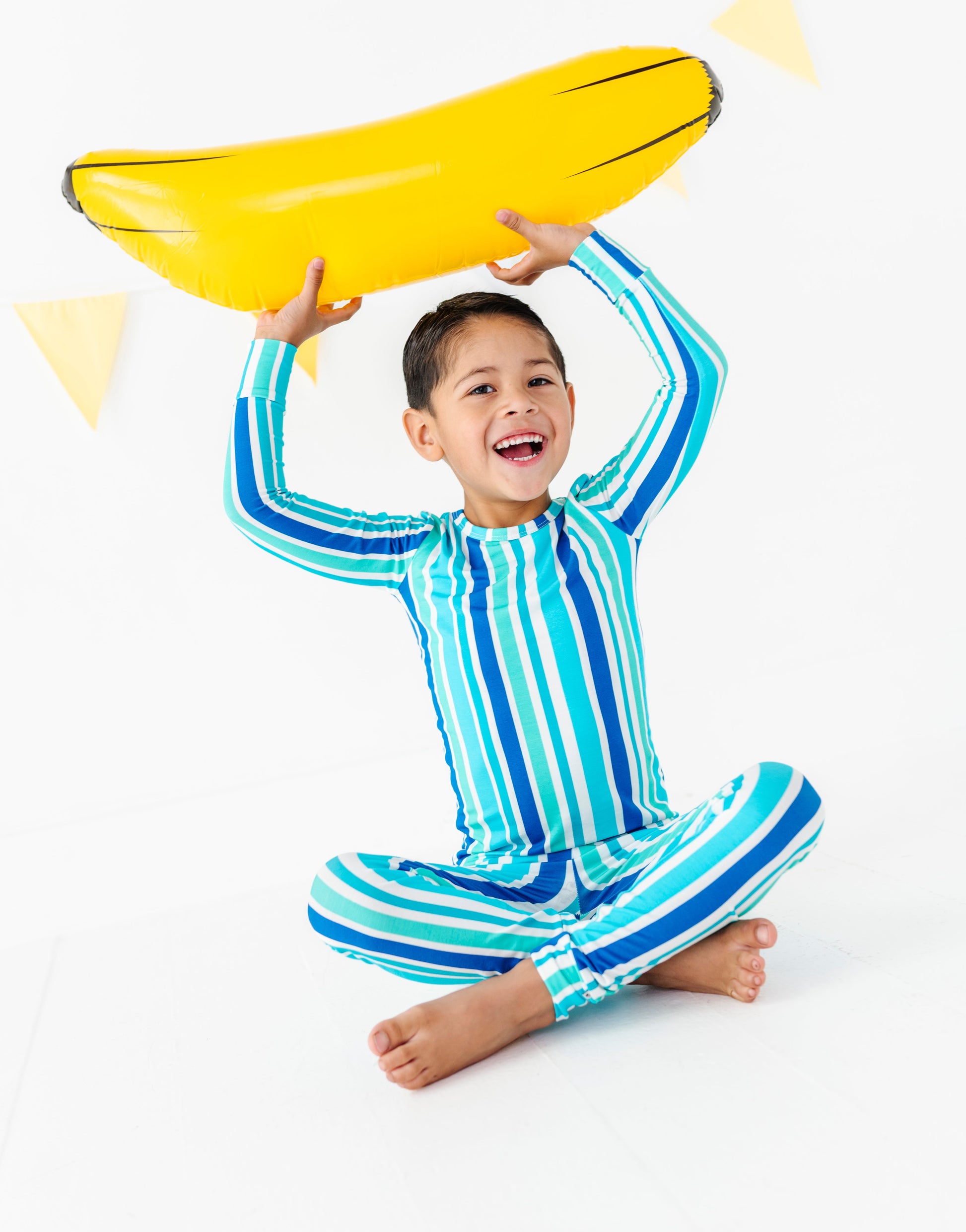 A young boy wearing a blue and green striped long-sleeve swim shirt and matching swim pants sits cross-legged on a white studio floor, holding a yellow inflatable banana-shaped float above his head.