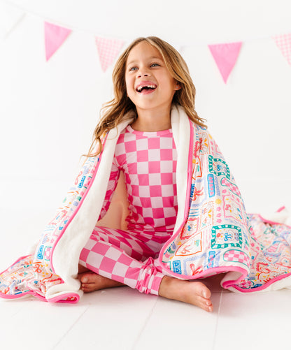 Child wrapped in a colorful blanket with a checkered outfit, sitting on a white surface with pink triangle flags in the background.