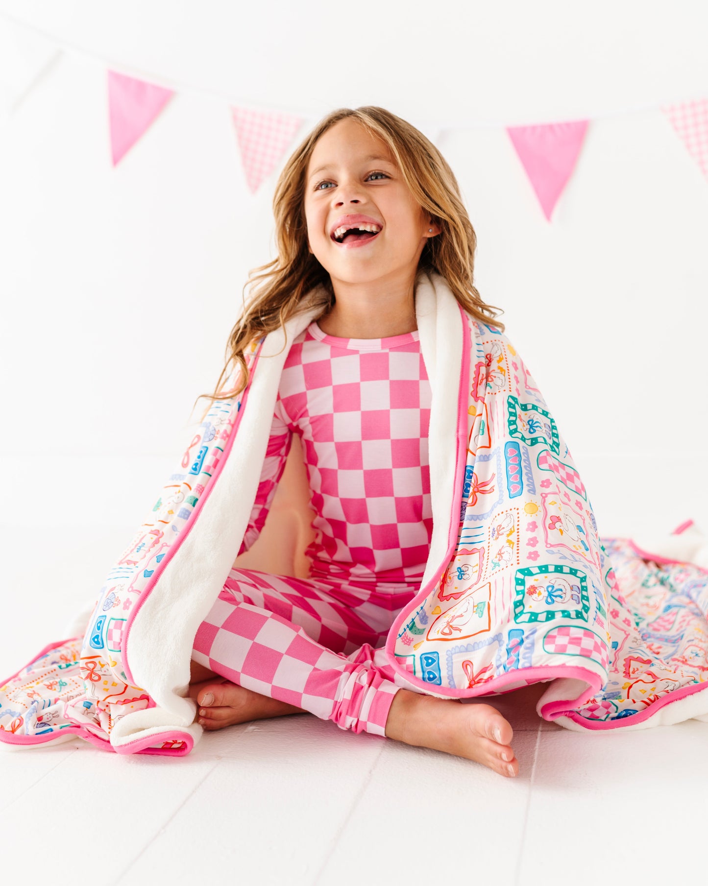 Child wrapped in a colorful blanket with  checkered pajamas, sitting on a white surface with pink triangle flags in the background.