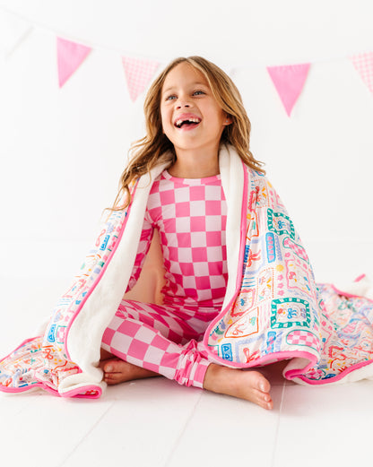 Child wrapped in a colorful blanket with  checkered pajamas, sitting on a white surface with pink triangle flags in the background.