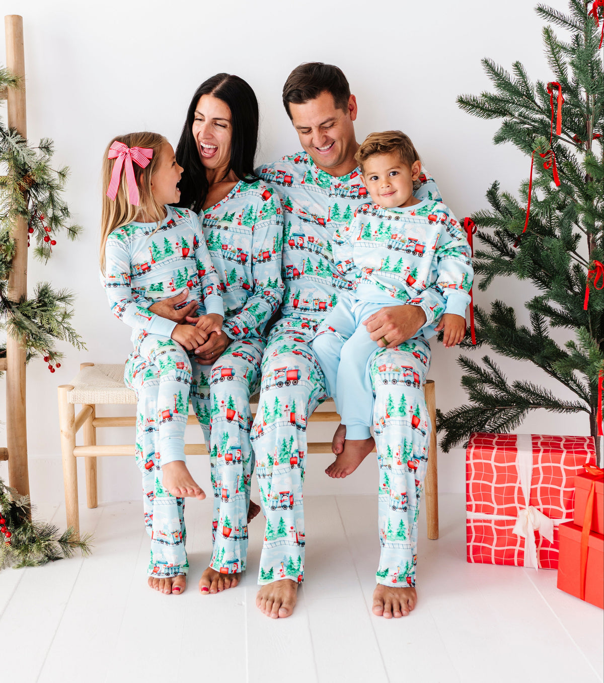 A family of four wearing matching light blue and white Christmas-themed pajamas, sitting together on a white wooden floor in front of a decorated Christmas tree and ladder shelf.
