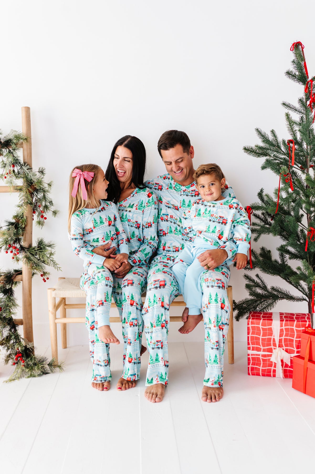 A family of four wearing matching light blue and white Christmas-themed bamboo pajamas, sitting together on a white wooden floor in front of a decorated Christmas tree and ladder shelf.