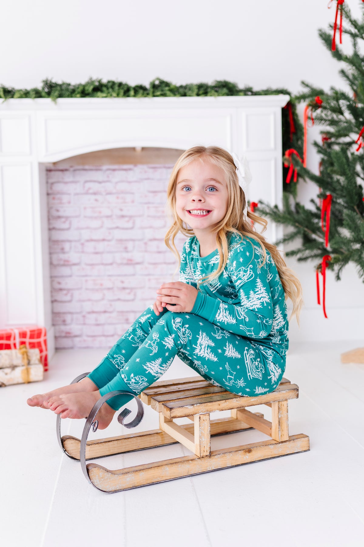 A little girl sitting on a wooden sleigh. She's wearing bamboo pajamas featuring a vintage print, including Santa, his reindeer, pine trees, and sleighs 