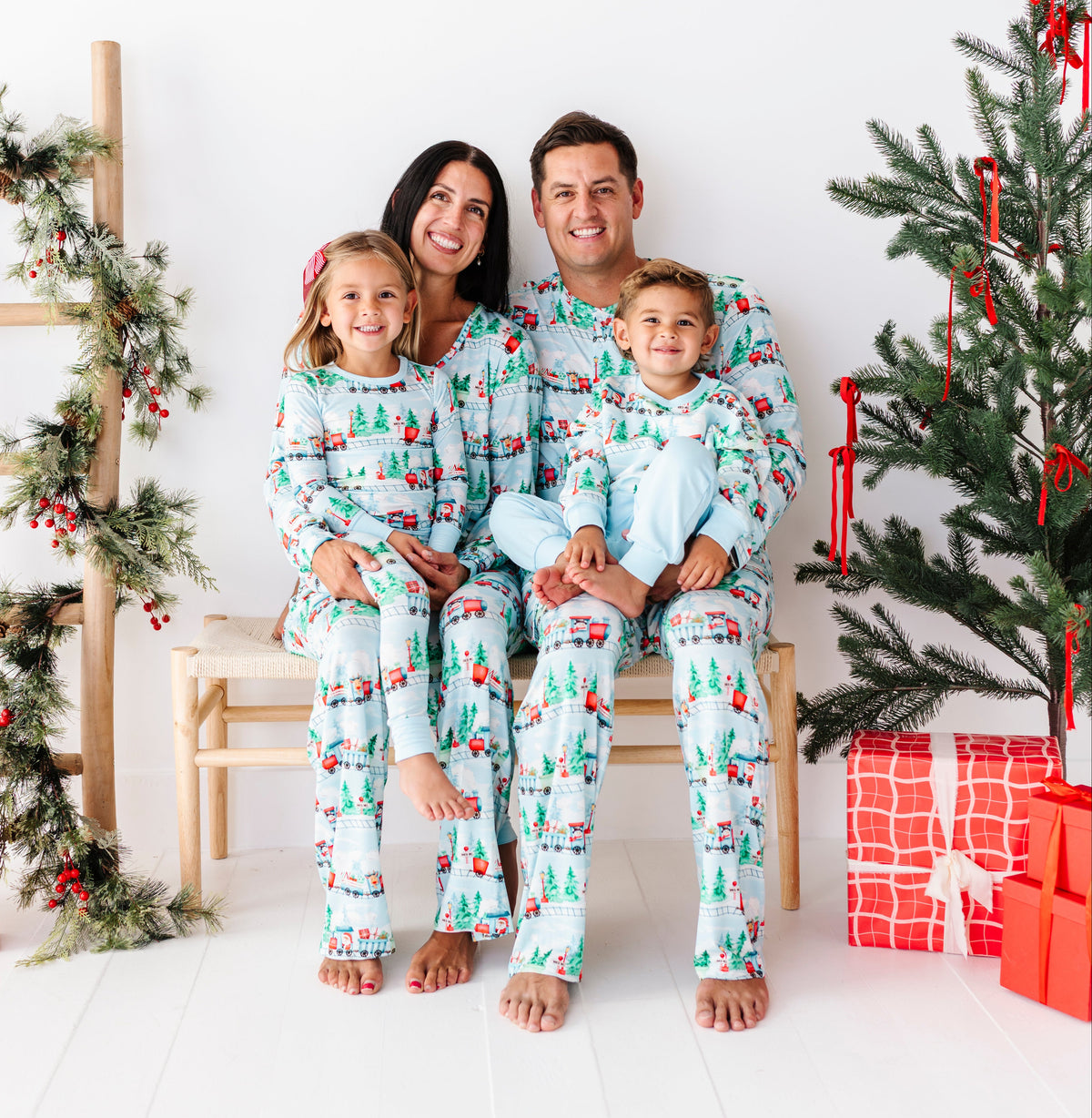 A family of four wearing matching light blue Christmas-themed pajamas sits together on a wooden bench in a bright, white holiday-themed room with a decorated Christmas tree and wrapped presents.