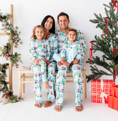 A family of four wearing matching light blue Christmas-themed pajamas sits together on a wooden bench in a bright, white holiday-themed room with a decorated Christmas tree and wrapped presents.