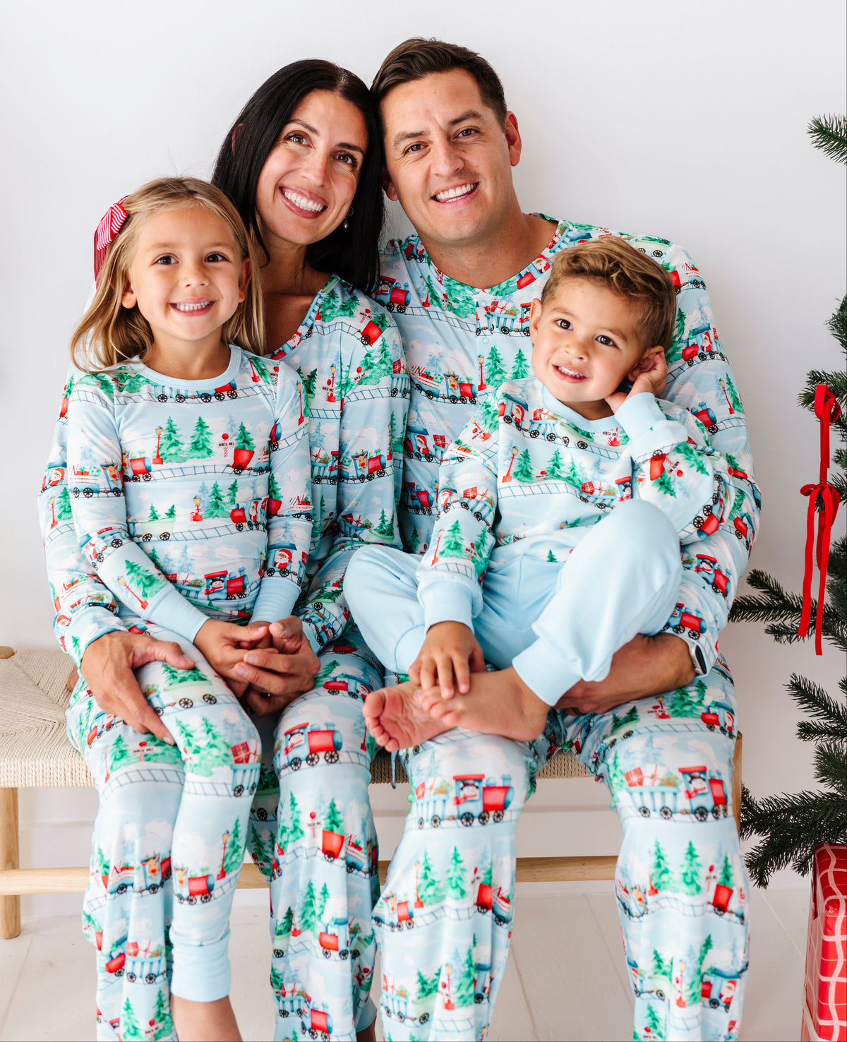A family of four wearing matching light blue Christmas-themed pajamas with a festive print of trees, sleighs, and red houses, posed together on a bench in front of a white wall.