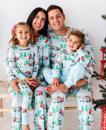 A family of four wearing matching light blue Christmas-themed pajamas with a festive print of trees, sleighs, and red houses, posed together on a bench in front of a white wall.