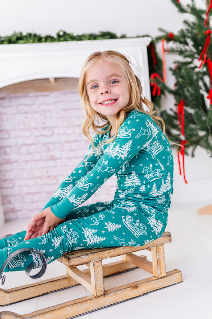A little girl sitting on a wooden sleigh. She's wearing bamboo pajamas featuring a vintage scene with Santa and his reindeer flying through the night sky 