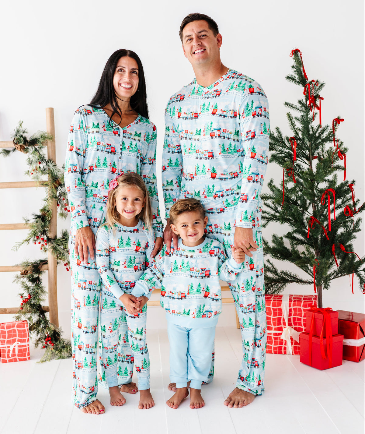 A family of four wearing matching Christmas-themed long-sleeve pajamas stands together in a bright studio setting with a decorated Christmas tree, ladder, and wrapped gifts.