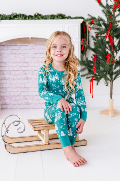 A blonde little girl wearing a bamboo two-piece pajama set. The pajamas feature vintage sleighs, Santas, reindeer, and pine trees. She's sitting on a wooden sleigh in front of a fireplace and a ribbon-filled Christmas tree
