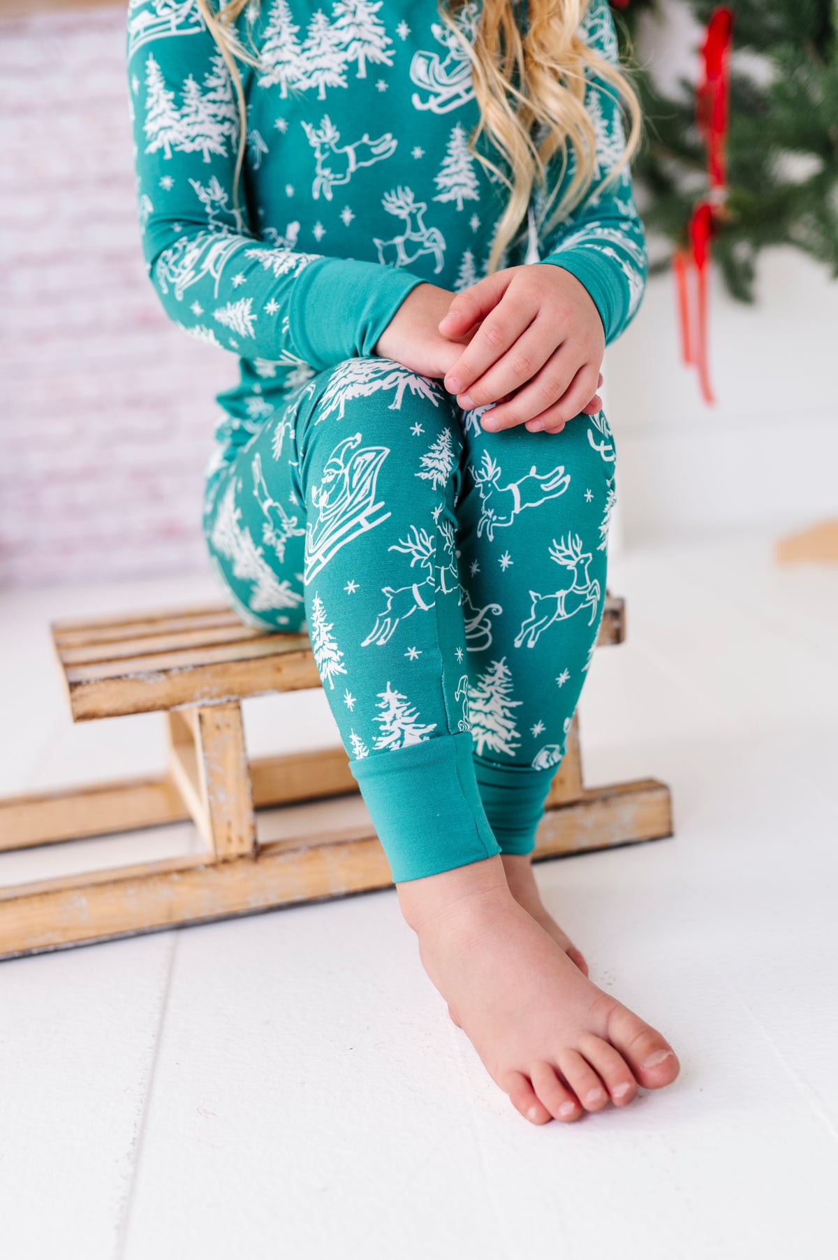 A close-up photo of a little girl wearing a bamboo two-piece pajama set. The pajamas feature vintage sleighs, Santas, reindeer, and pine trees. She's standing in front of a fireplace and a ribbon-filled Christmas tree