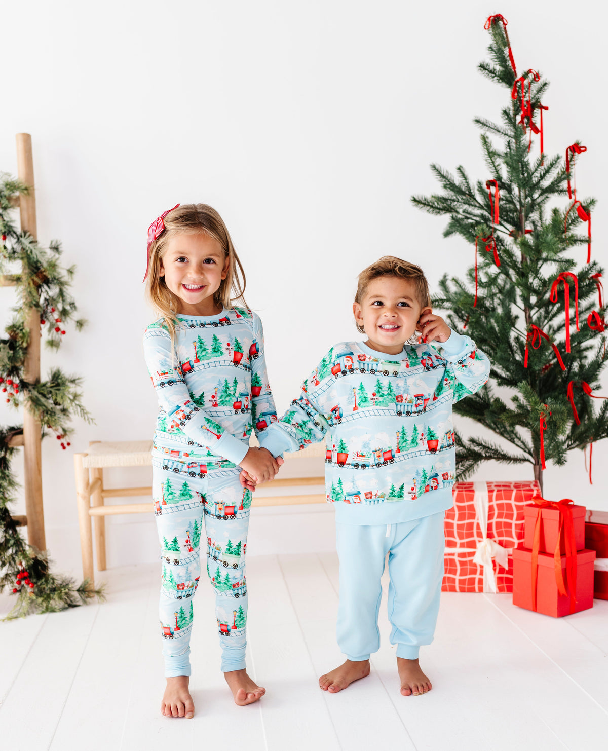A young girl and boy wearing matching light blue Christmas-themed bamboo pajamas stand holding hands in a bright white room decorated with a Christmas tree and wrapped presents.