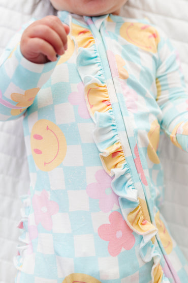 A close-up of a baby wearing a light blue and white checkered sleep sack with pastel smiley faces, pink flowers, and yellow ruffle trim along the front zipper.