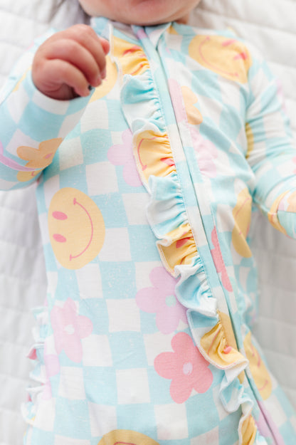 A close-up of a baby wearing a light blue and white checkered sleep sack with pastel smiley faces, pink flowers, and yellow ruffle trim along the front zipper.