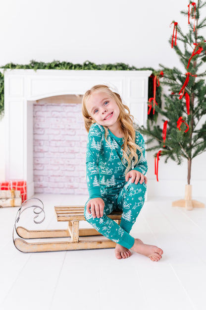 A blonde little girl wearing a bamboo two-piece pajama set. The pajamas feature vintage sleighs, Santas, reindeer, and pine trees. She's sitting on a wooden sleigh in front of a fireplace and a ribbon-filled Christmas tree