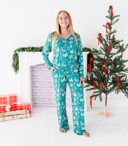 A blonde woman standing in front of a Christmas tree and a fireplace wearing bamboo pajamas. The super soft pajamas feature vintage Santa, his reindeer, and pine trees 