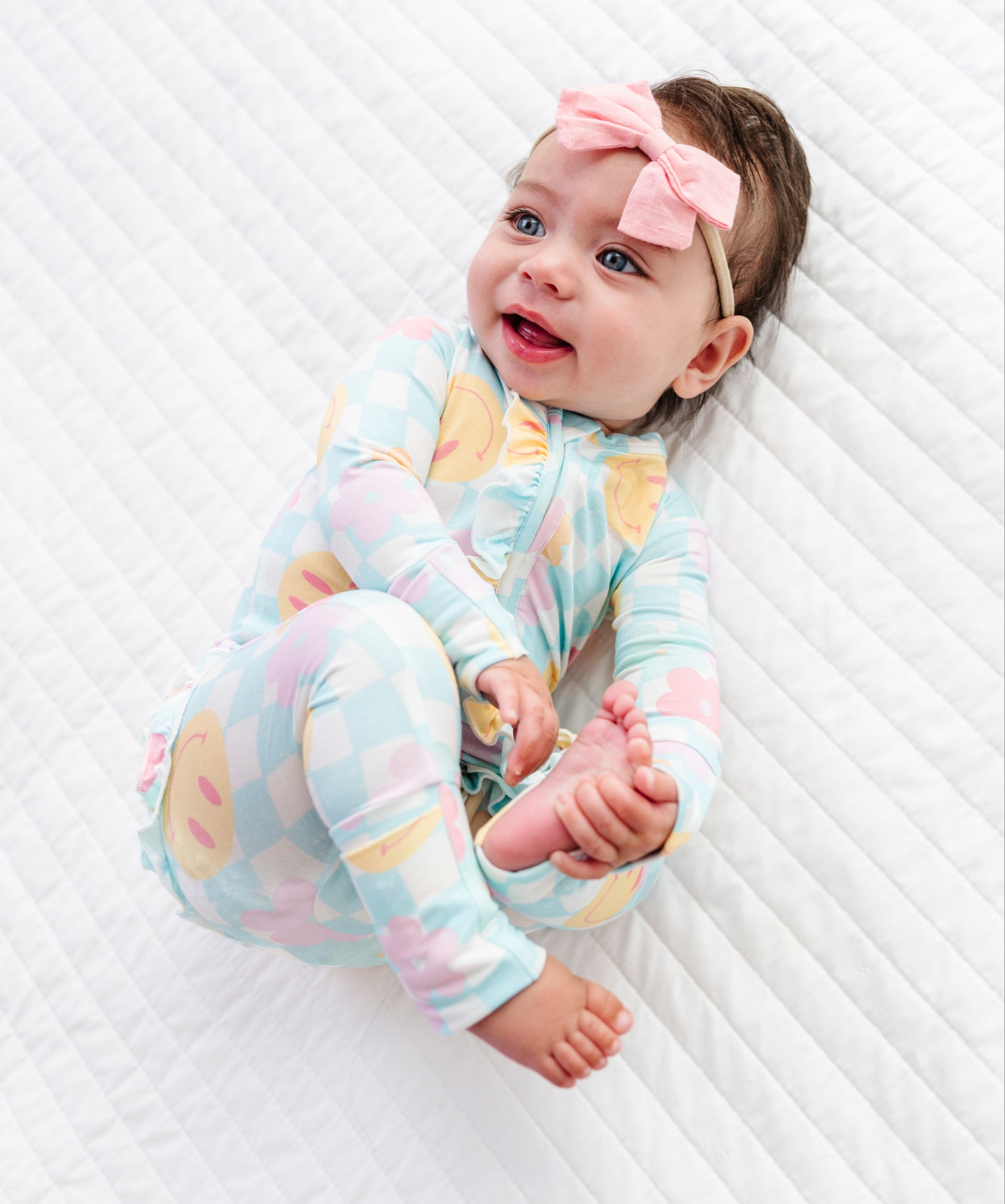 A baby lying on a white quilted mattress, wearing a pastel multicolor patterned long-sleeve footless onesie and a pink bow headband.