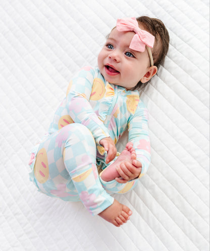 A baby lying on a white quilted mattress, wearing a pastel multicolor patterned long-sleeve footless onesie and a pink bow headband.