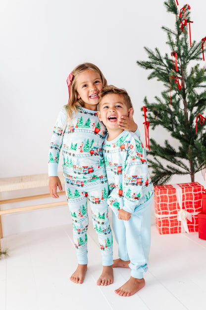 Two children wearing matching Christmas-themed bamboo pajamas standing together in a bright room next to a decorated Christmas tree and wrapped presents.