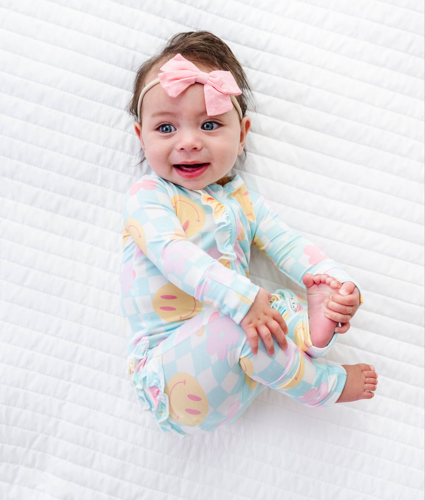 A baby lying on a white quilted blanket, wearing a pastel multicolor checkered long-sleeve pajama top with a pink bow headband.