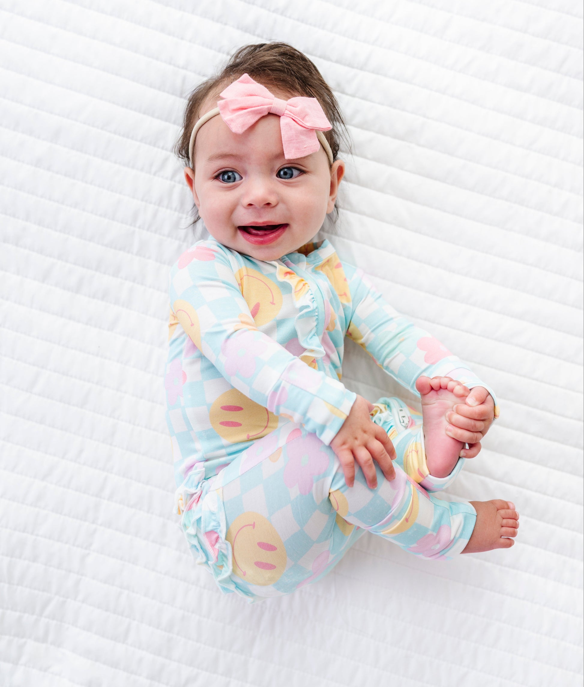 A baby lying on a white quilted blanket, wearing a pastel multicolor checkered long-sleeve pajama top with a pink bow headband.