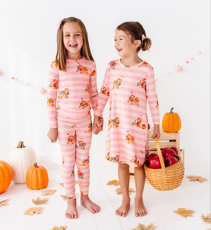 Two young girls standing barefoot and holding hands in a bright white studio decorated with pumpkins, fall leaves, and a basket of red apples. The girl on the left wears a pink long-sleeve pajama set with a cartoon character print, and the girl on the right wears a matching pink striped pajama dress with the same character print.